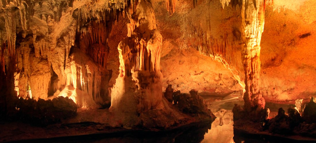 Beeindruckende Tropfsteinhöhle mit spektakulären Stalaktiten und Stalagmiten in warmen Rottönen. Die Spiegelung der Felsformationen im Wasser verstärkt die mystische Stimmung.