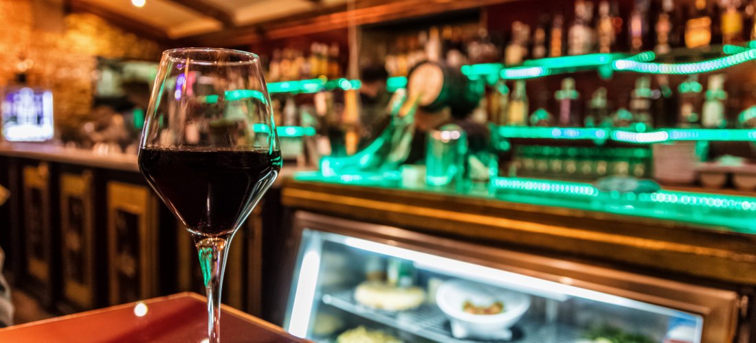 Nahaufnahme eines Glases Rotwein auf einem Tisch in einer Bar mit beleuchteten Regalen und Theke im Hintergrund