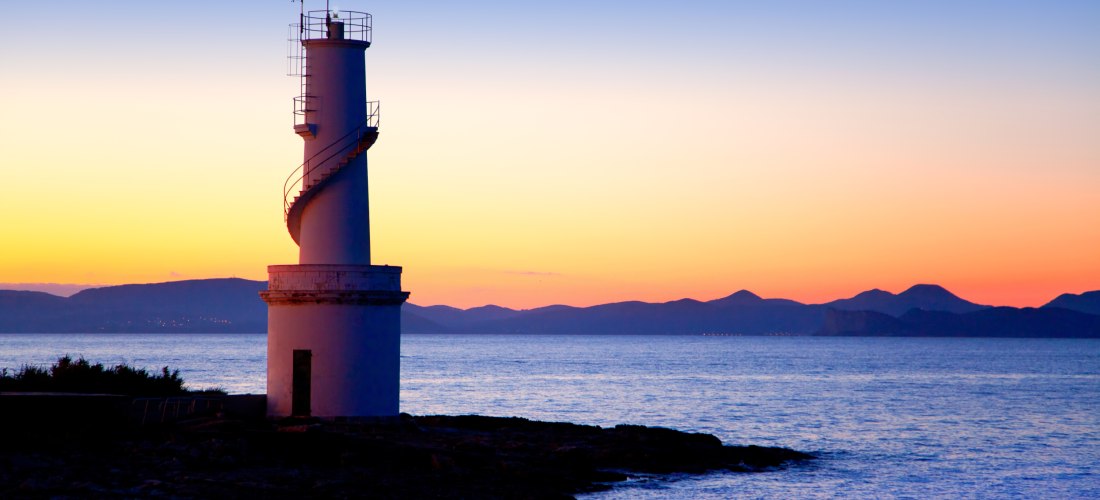 Weißer Leuchtturm am Meer mit spiralförmiger Außentreppe, aufgenommen bei Sonnenuntergang. Der Himmel leuchtet in kräftigen Orangetönen, im Hintergrund sind die Umrisse von Bergen zu sehen.