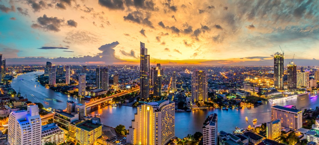 Atemberaubende Panoramaaufnahme der Skyline von Bangkok bei Sonnenuntergang. Der Fluss Chao Phraya schlängelt sich durch die beleuchtete Stadt, gesäumt von modernen Hochhäusern und Brücken. Der Himmel leuchtet in dramatischen Orange-, Gelb- und Blautönen 