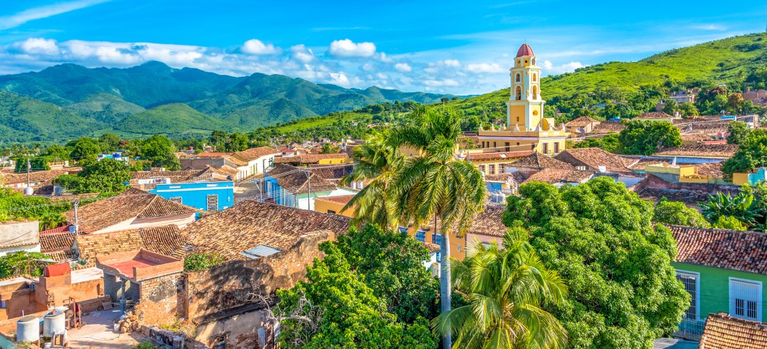 Trinidad auf Kuba – Koloniale Altstadt mit farbenfrohen Häusern und Blick auf die Berge Panoramablick über die historische Altstadt von Trinidad auf Kuba mit farbenfrohen Kolonialhäusern, Palmen, dem gelben Glockenturm der Kirche und der Sierra del Escambray im Hintergrund.