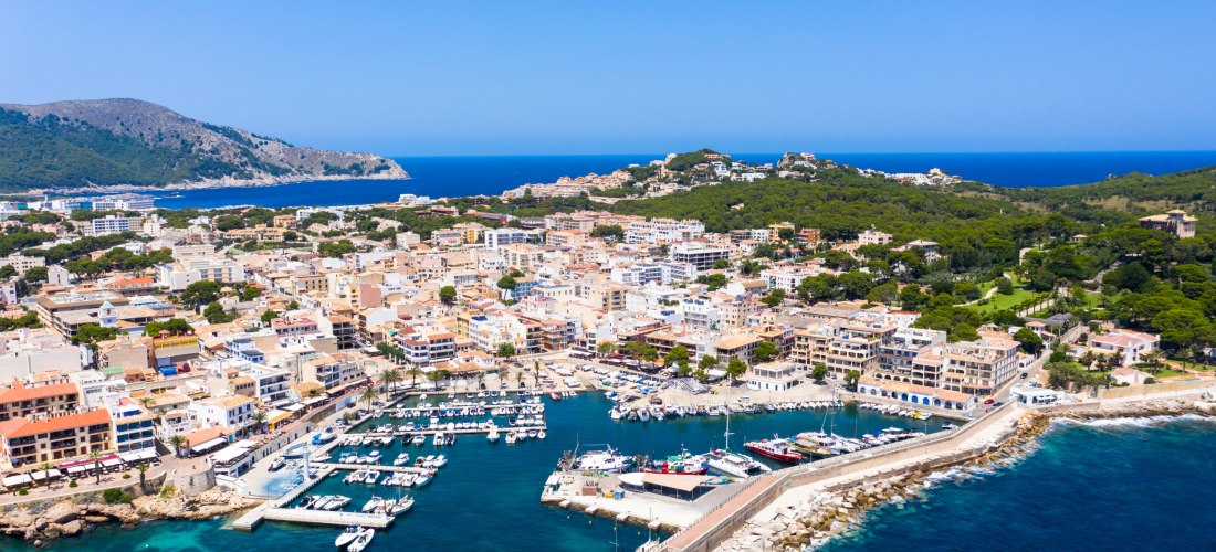 Luftaufnahme der Stadt Cala Ratjada mit Blick auf den Hafen, in dem zahlreiche Boote und Yachten liegen. Die Stadt ist dicht bebaut mit hellen, mediterranen Häusern, teils mit Flachdächern und Dachterrassen. Eine Promenade verläuft entlang des Wassers, ge