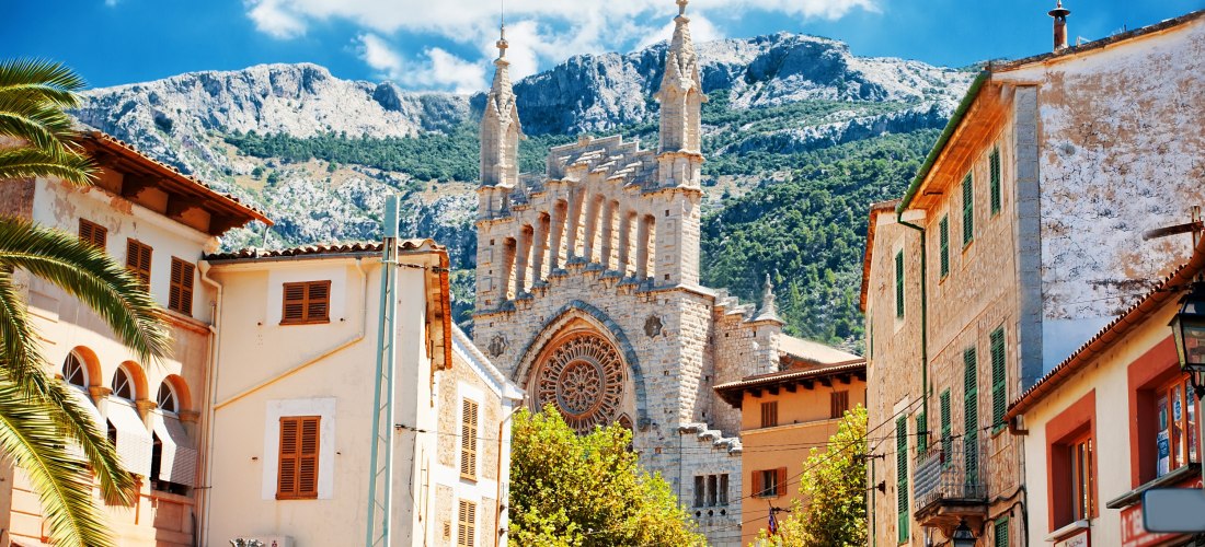 Eine gotische Kirche auf Mallorca mit einer imposanten Fassade und detaillierten Steinmetzarbeiten. Im Vordergrund sind Gebäude mit typisch mediterraner Architektur zu sehen, darunter Fassaden mit grünen Fensterläden. Im Hintergrund ragen Berge auf, die d