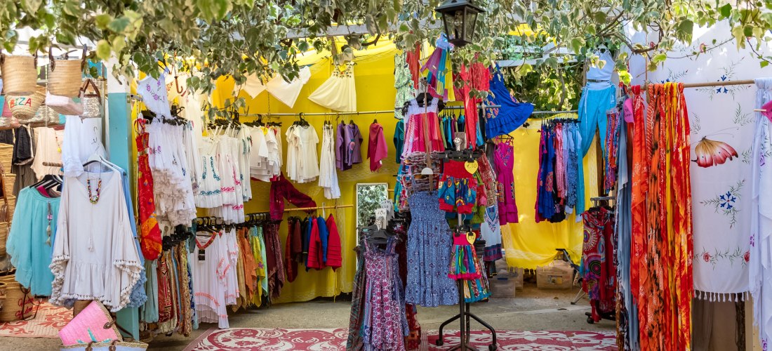 Bunter Hippiemarkt auf Ibiza: Unter einem Blätterdach hängen und liegen farbenfrohe Kleidungsstücke wie Kleider, Tuniken und Tücher. An einem Kleiderständer sind Sommerkleider in kräftigen Farben ausgestellt. Die Marktstände sind mit gelben Stoffbahnen de