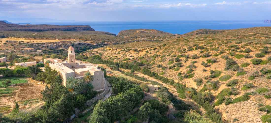 Historisches Kloster mit Glockenturm inmitten einer trockenen, hügeligen Landschaft mit spärlicher Vegetation. Im Hintergrund ist das Meer unter leicht bewölktem Himmel sichtbar