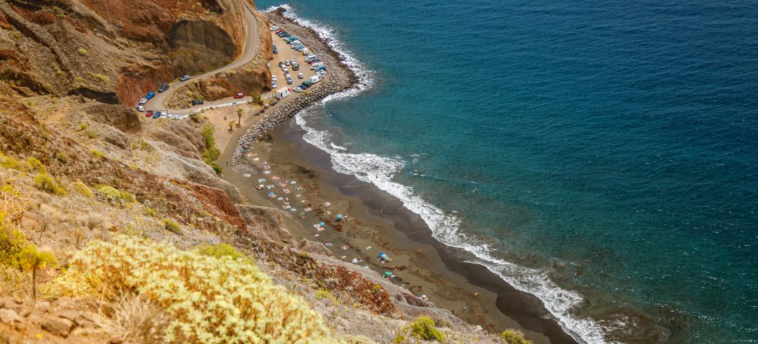 abgelegener Naturstrand mit dunklem Sand, umgeben von Felsen und Meer, erreichbar per Boot oder Wanderung.