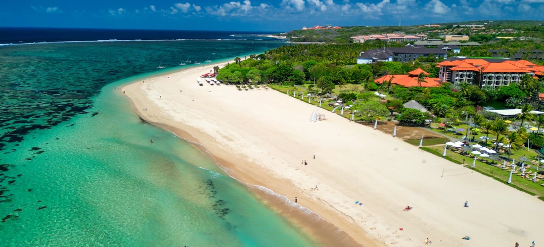 Breiter Sandstrand von Nusa Dua auf Bali mit türkisblauem Wasser, grünen Palmen und roten Dächern der Hotelanlagen im Hintergrund