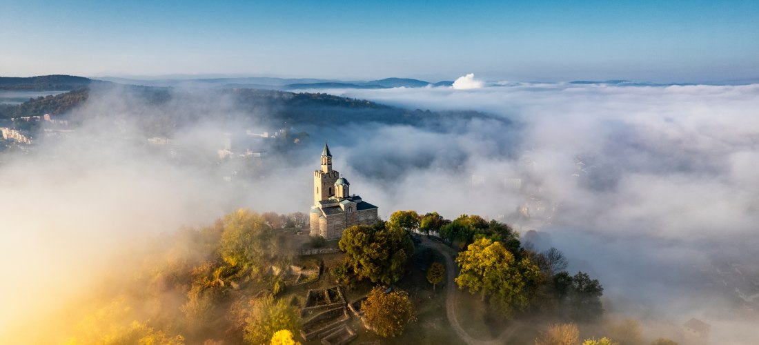 Alte Burg auf einem Hügel umgeben von buntem Herbstlaub, eingetaucht in dichte Nebelschwaden, mit klar blauem Himmel darüber