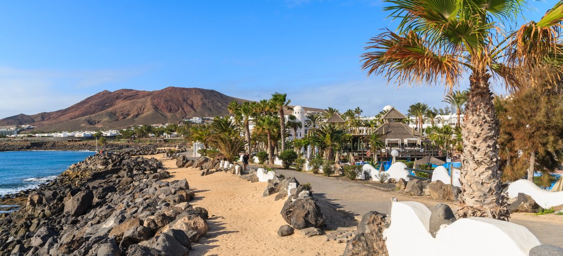 Promenade von Playa Blanca auf Lanzarote mit Palmen, Vulkangebirge im Hintergrund und Blick auf das Meer.