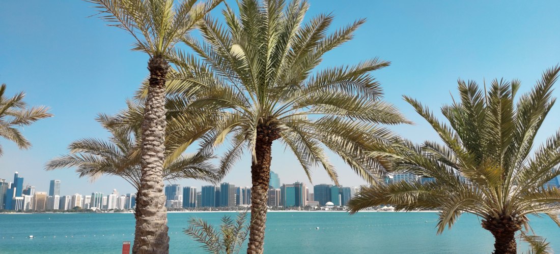 Palmen am weißen Sandstrand mit türkisfarbenem Wasser, im Hintergrund die moderne Skyline von Abu Dhabi