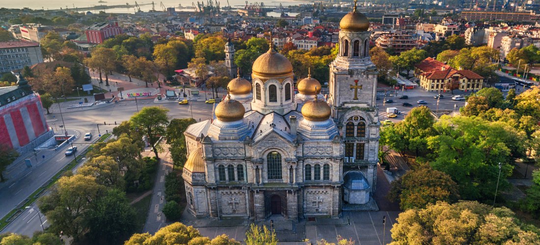 Luftaufnahme der Kathedrale von Varna in Bulgarien. Das imposante Bauwerk besitzt goldene Zwiebeltürme und ist umgeben von Bäumen und Straßen. Im Hintergrund die Stadt Varna, ein Hafen und das Schwarze Meer unter leicht bewölktem Himmel