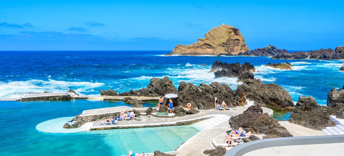 Natürliche Lavapools in Porto Moniz auf Madeira mit kristallklarem, türkisfarbenem Wasser, umgeben von schwarzen Vulkanfelsen. Menschen entspannen auf Sonnenliegen unter blauem Himmel mit Blick auf das offene Meer.