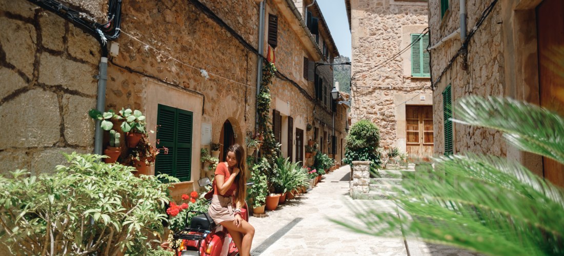 Enge, charmante Gasse in einem mallorquinischen Dorf mit Natursteinfassaden und grünen Fensterläden. Die Straße ist gesäumt von zahlreichen Blumentöpfen mit roten Geranien und üppigem Grün. Eine junge Frau in sommerlicher Kleidung sitzt lächelnd auf einem