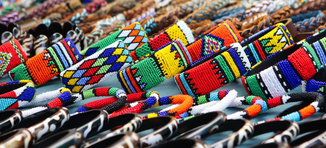 Nahaufnahme eines Marktstandes mit vielen farbenfrohen, handgefertigten Armbändern aus Perlen in traditionellen Mustern und Farben. Im Vordergrund sind auch Schnitzarmbänder zu sehen.