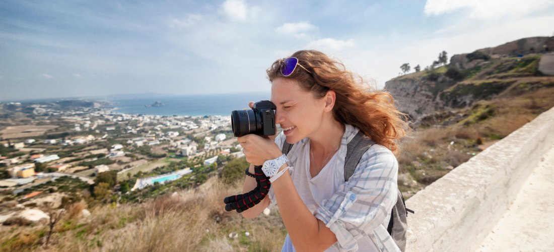 Junge Frau mit Kamera am Auge fotografiert eine weite Landschaft auf Kos. Im Hintergrund liegt eine weite Küstenlandschaft mit Häusern, Feldern und dem Meer. Die Frau trägt Freizeitkleidung, hat windzerzaustes Haar und wirkt konzentriert und glücklich.