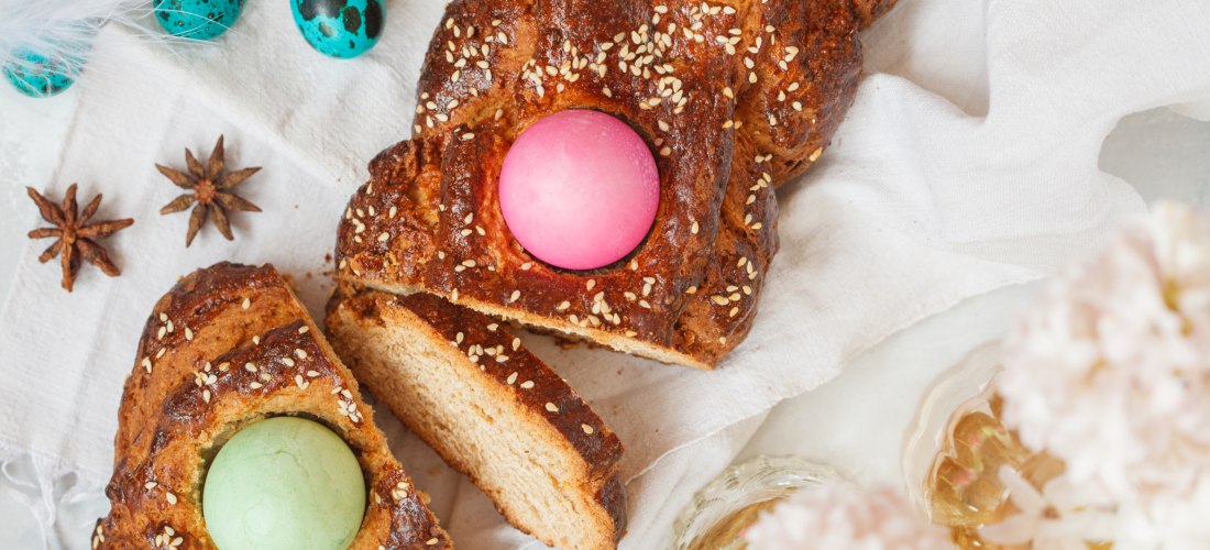Griechisches Osterbrot, verziert mit Sesamkörnern und eingefassten bunten Ostereiern in Pink und Grün. Das Brot liegt auf einem weißen Tuch, daneben dekorative blaue Wachteleier, Sternanis und zwei Gläser mit Blumen.