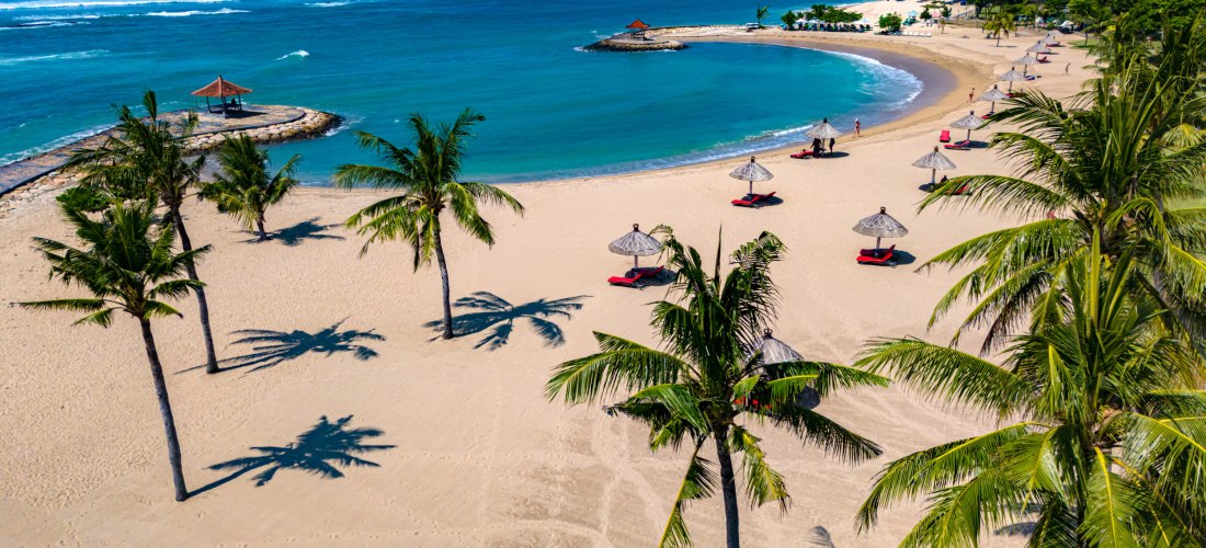 Tropischer Strand in Nusa Dua, Bali, mit Palmen, weißen Sonnenschirmen und ruhigem türkisblauen Meer