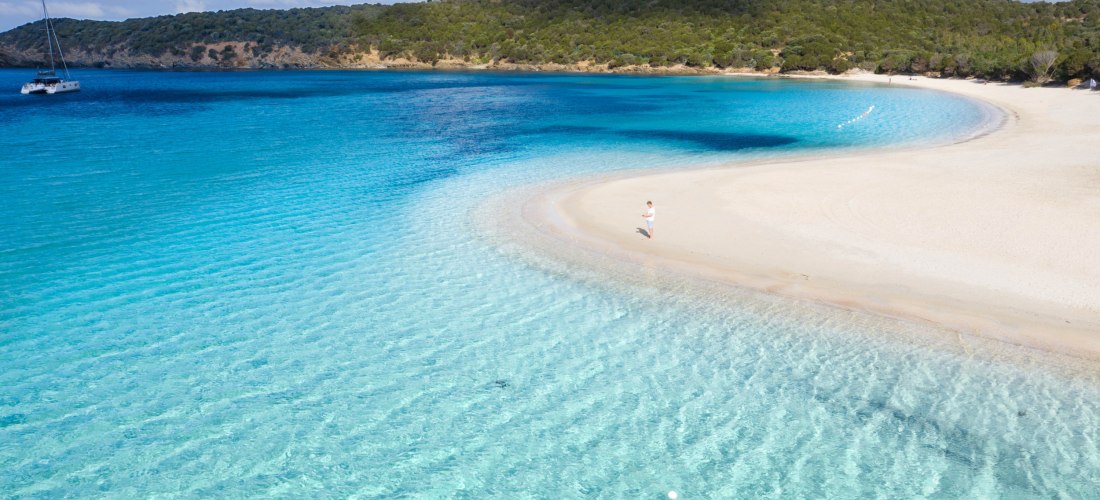 Heller Sandstrand mit seichtem, türkisblauem Wasser. Eine einzelne Person steht am Ufer, im Hintergrund dichter Wald und ein Boot.