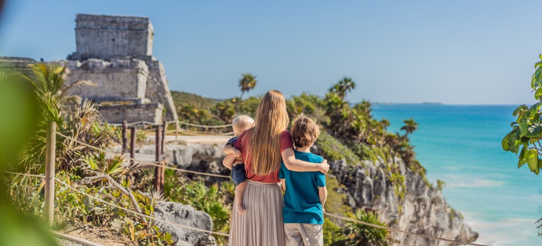 Mutter mit zwei Kindern betrachtet die alte Maya-Ruine an einer Klippe mit Blick auf das türkisfarbene Meer, umgeben von tropischer Vegetation