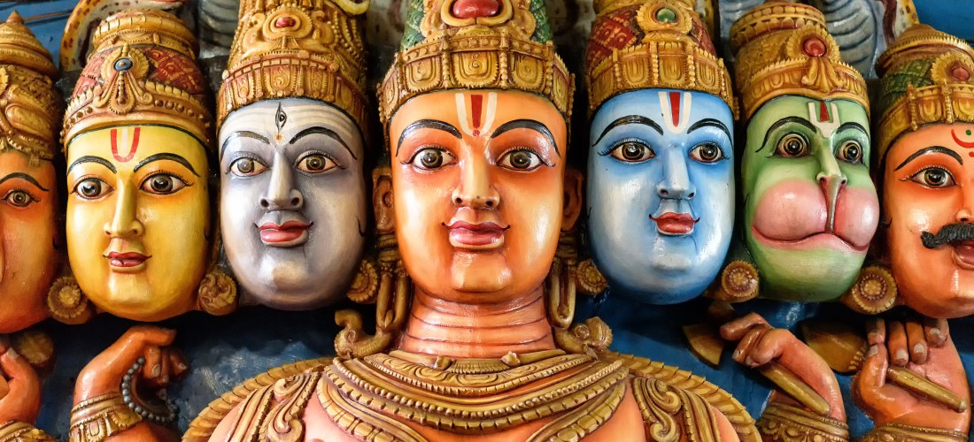 Detailaufnahme einer farbenfrohen Götterstatue mit mehreren Gesichtern aus einem hinduistischen Tempel. Die Köpfe sind kunstvoll bemalt in verschiedenen Farben wie Orange, Blau, Grün und Gold, geschmückt mit Kronen und Schmuck. Im Hintergrund sind mehrere