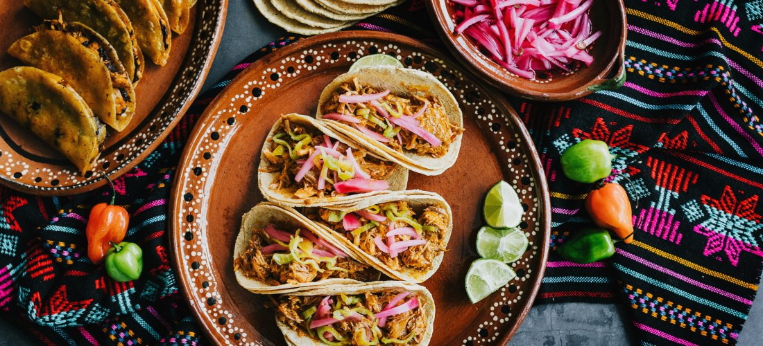 Traditionelle mexikanische Tacos mit Fleisch, eingelegten Zwiebeln und Limetten auf einem bemalten Keramikteller. Daneben Mais-Tortillas, Chili, rote Zwiebeln und bunte Stoffe mit folkloristischen Mustern. Das Bild vermittelt Genuss, Kultur und landestypi