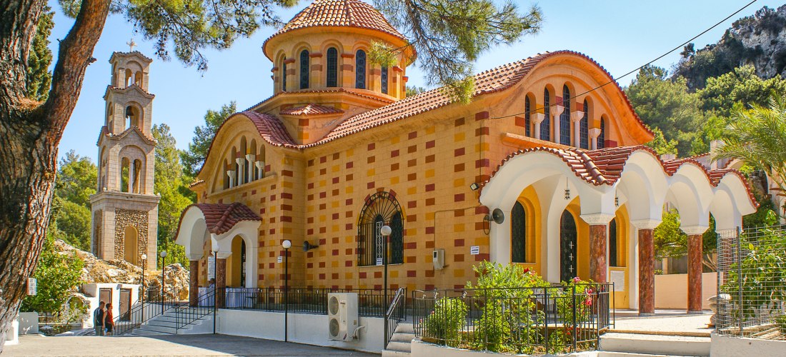 Orthodoxe Kirche in einem Pinienhain auf Rhodos: Die farbenfrohe Fassade der Kirche mit ihrem wellenförmigen Vordach und dem markanten Kuppeldach strahlt in der Sonne. Der danebenstehende Glockenturm wirkt filigran. Die Umgebung ist ruhig, umgeben von Nat