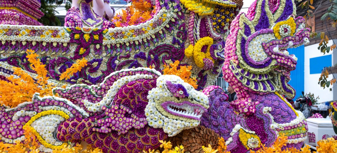Prächtig geschmückter Umzugswagen mit traditionellen Figuren, komplett aus bunten Blüten gestaltet. Farben wie Lila, Gelb, Orange und Weiß dominieren. Dargestellt sind Drachen und mythologische Motive, umgeben von Orchideen.