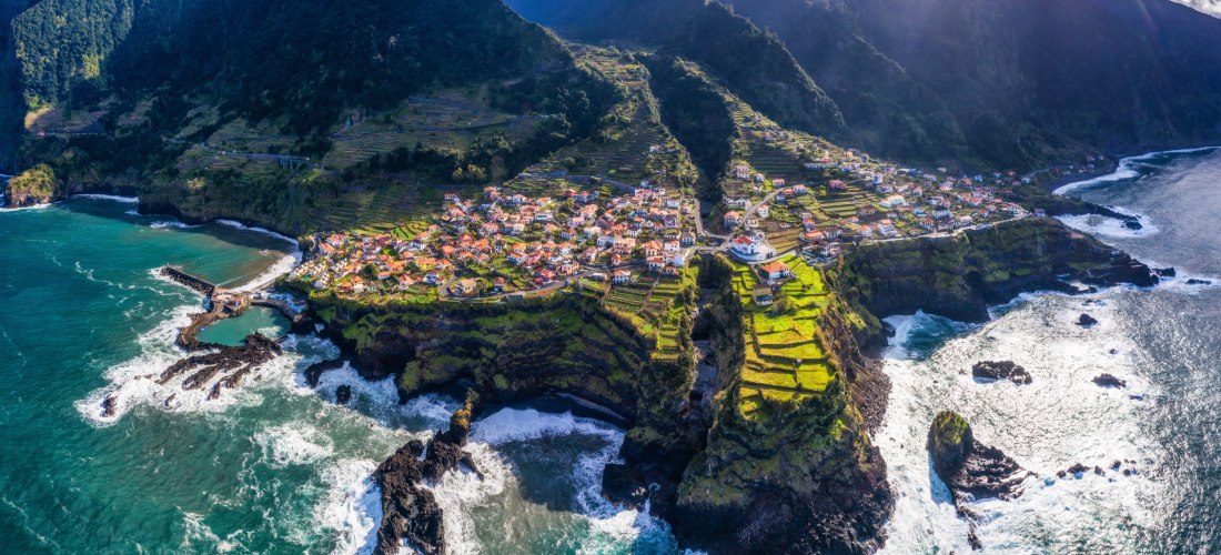 Das Foto zeigt Felsterrassen mit kleinen Häusern und Feldern, die steil bis zum Meer abfallen.      Zwischen Lavafelsen bricht sich die kräftige Brandung; Berge bilden die dunkle, wolkenumspielte Kulisse.