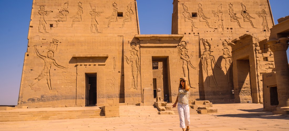 Ägypten - Assuan Das Bild zeigt eine Frau, die vor dem monumentalen Eingang des Philae-Tempels nahe Assuan steht. Die Tempelfassade ist mit präzise gemeißelten Reliefs geschmückt, die Szenen aus der altägyptischen Mythologie zeigen – darunter Götterfiguren wie Isis, Horus