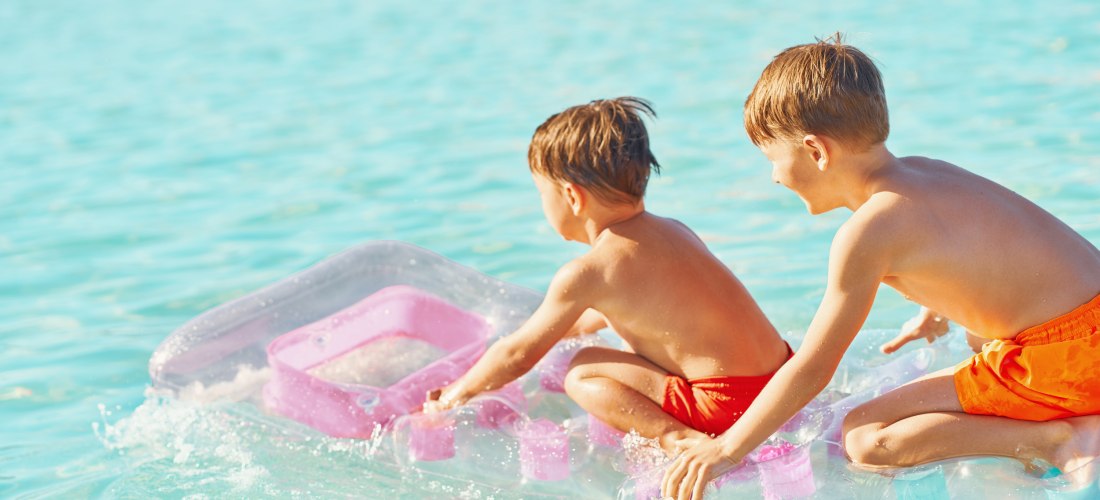 Zwei Kinder in orangefarbener Badebekleidung knien fröhlich auf einer pink-transparenten Luftmatratze im flachen, türkisblauen Meer. Sie lachen, plantschen und genießen das sonnige Badevergnügen.
