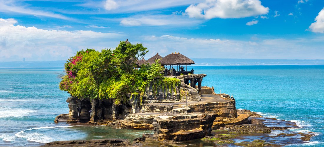 Der Tempel Tanah Lot auf Bali steht malerisch auf einem Felsen im Meer, umgeben von türkisfarbenem Wasser. Der Tempel besteht aus mehreren Pavillons mit traditionellen, dunklen Strohdächern und ist über eine steinerne Treppe erreichbar. Üppiges, tropische