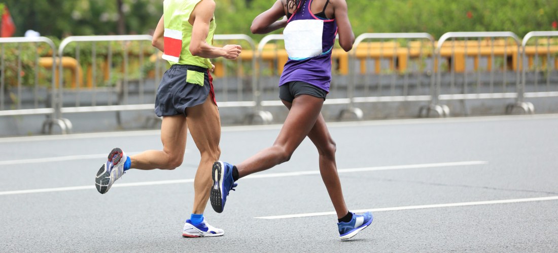 Zwei Marathonläufer:innen in Sportkleidung laufen nebeneinander auf einer abgesperrten Straße. Der Fokus liegt auf ihren Beinen in Bewegung, im Hintergrund sind Absperrgitter und Grünanlagen zu sehen.
