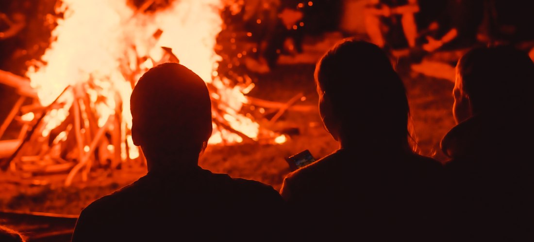 Menschen sitzen im Dunkeln um ein großes Lagerfeuer, das hell lodert. Die Flammen werfen ein warmes Licht auf die Umstehenden, eine Person hält ein Handy in der Hand.