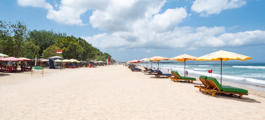 Weißer Sandstrand mit bunten Sonnenschirmen und Sonnenliegen am Kuta Beach auf Bali