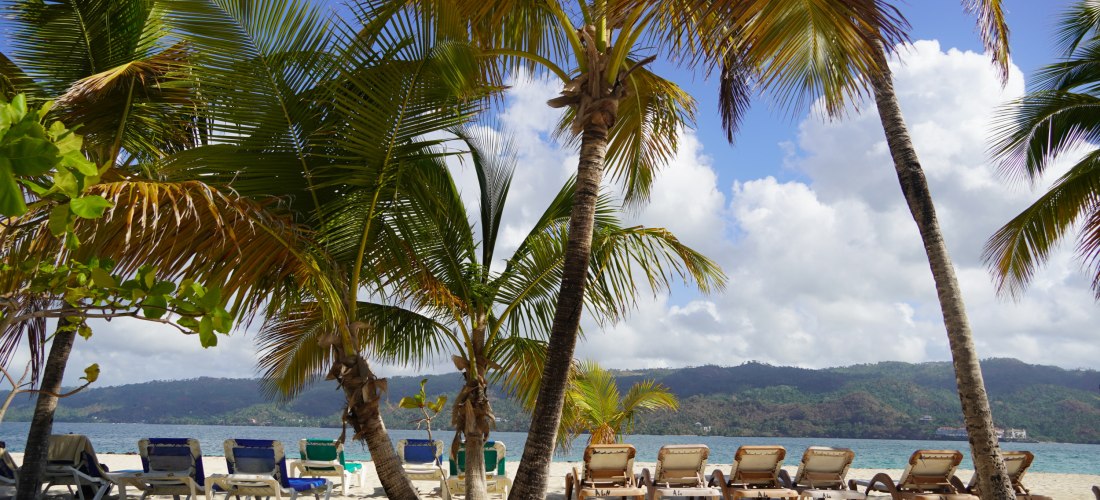 Sonnenliegen im Schatten von Palmen am Strand von Samaná in der Dominikanischen Republik mit Blick auf das Meer und die grünen Hügel im Hintergrund