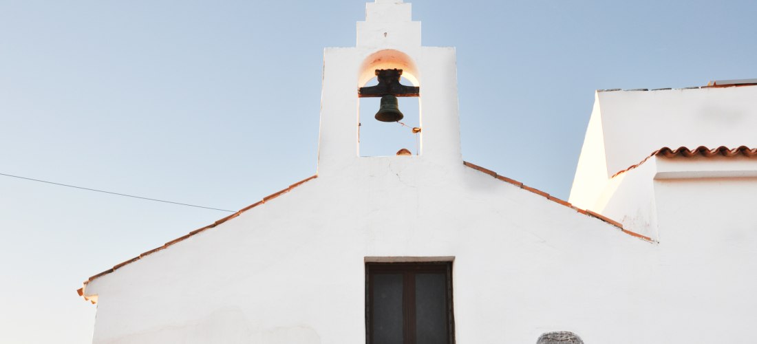 Eine kleine weiße Kirche mit einfacher Fassade, einem dunklen Fenster und einem Glockengiebel mit Kreuz auf dem Dach. Die Glocke hängt frei sichtbar unter dem blauen Himmel. Die Architektur wirkt minimalistisch und traditionell, typisch für den Mittelmeer