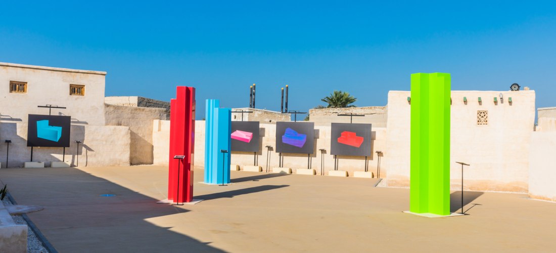 Moderne Freiluft-Kunstausstellung mit bunten Skulpturen in Rot, Blau und Grün, umgeben von traditioneller sandfarbener Architektur
