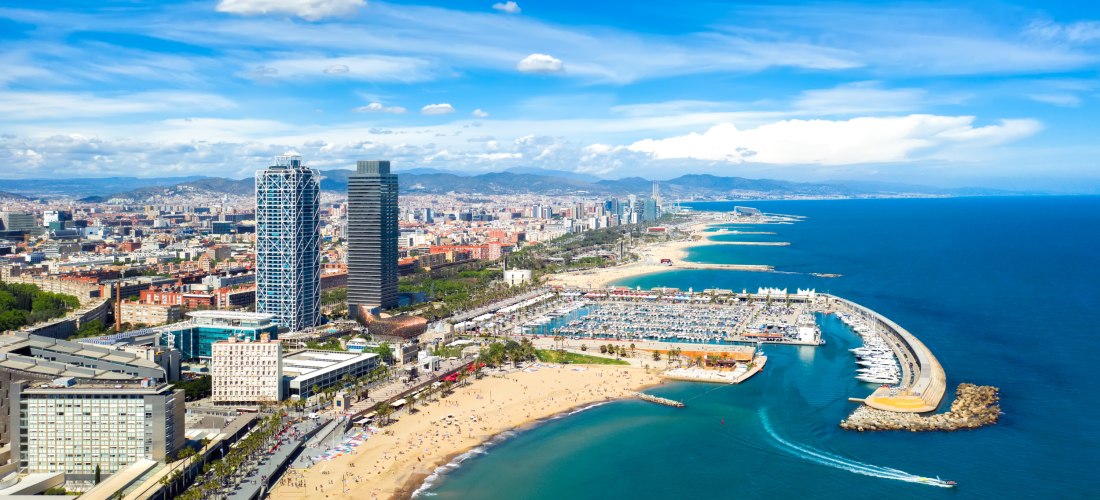 Luftaufnahme von Barcelona mit Blick auf die Küste, den langen Sandstrand, die Marina Port Olímpic und die markanten Hochhäuser Torre Mapfre und Hotel Arts. Im Hintergrund die Stadt und umliegende Berge unter blauem Himmel.