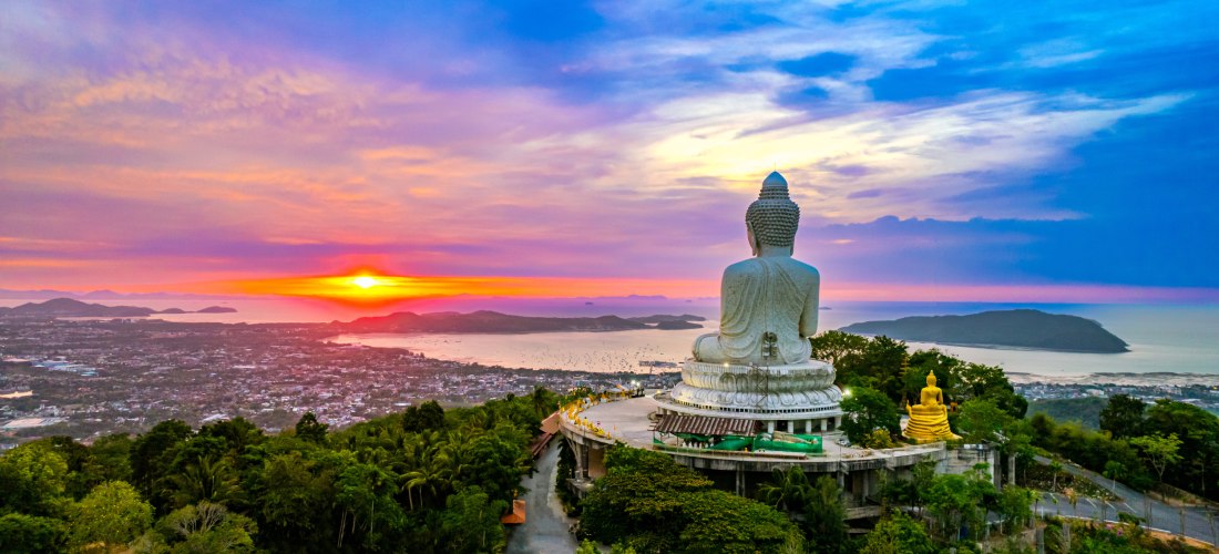 Panoramablick auf die berühmte weiße Buddha-Statue auf dem Nakkerd Hill in Phuket, Thailand. Die Statue blickt auf die Stadt und das Meer hinaus, während im Hintergrund die Sonne spektakulär untergeht. Der Himmel ist in kräftigen Farben gefärbt – von Oran