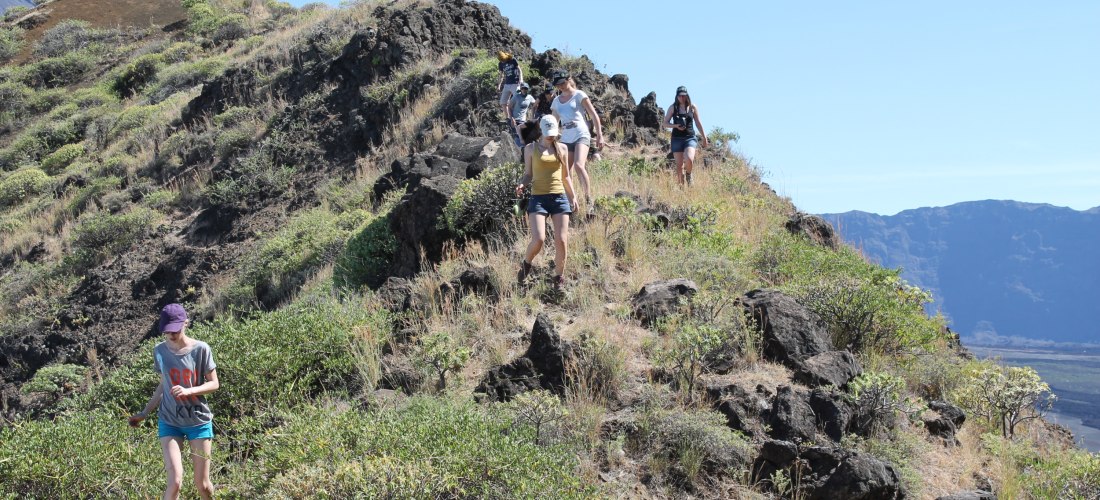 Eine Gruppe von Wandernden steigt einen schmalen, felsigen Pfad in einer hügeligen, trockenen Landschaft mit niedriger Vegetation hinab. Die Szene wirkt sonnig und offen, im Hintergrund sind steile Felswände zu sehen.