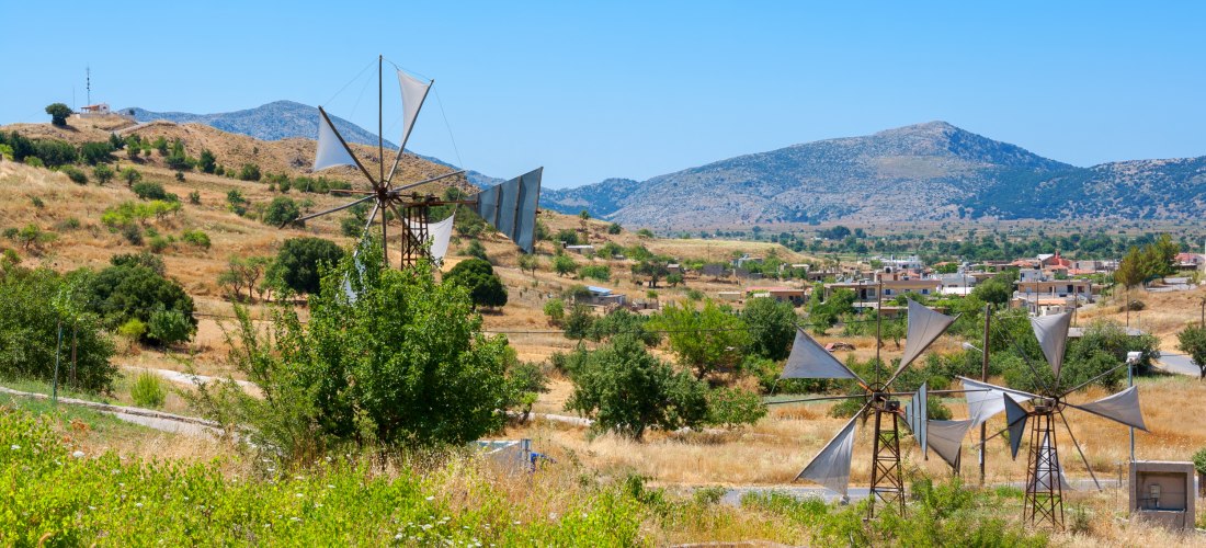 Traditionelle Windmühlen mit weißen Segeltuchflügeln stehen in einer trockenen, hügeligen Landschaft mit vereinzelten Bäumen. Im Hintergrund sind ein kleines Dorf und Berge unter blauem Himmel zu sehen