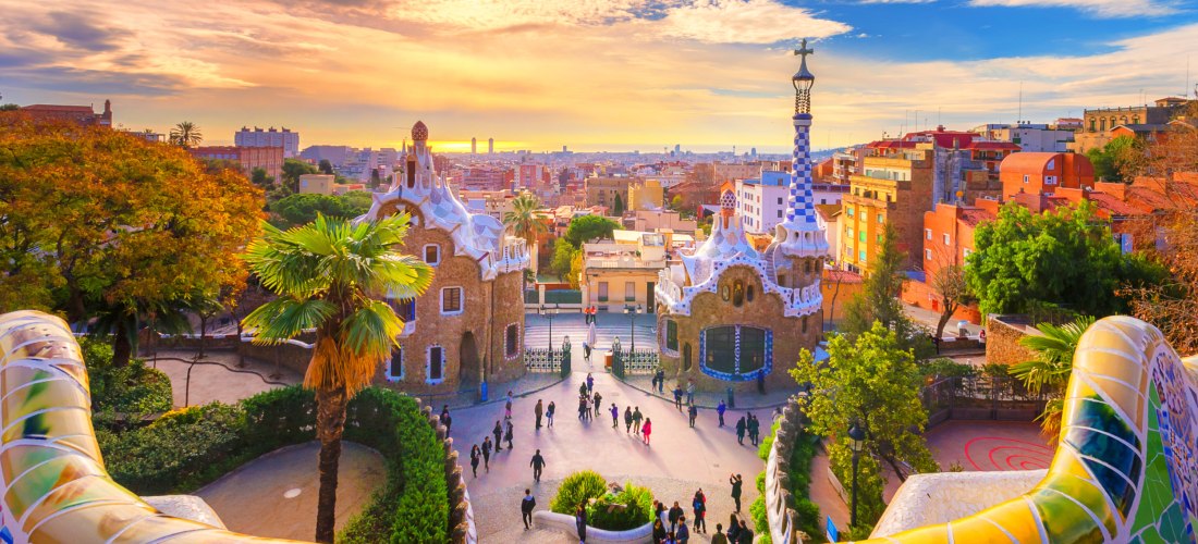 Blick vom Park Güell in Barcelona mit seinen berühmten, bunten Mosaikbänken auf die Stadt. Zwei märchenhafte Pavillons im Vordergrund, dahinter die Dächer der Stadt bei goldenem Sonnenuntergang unter einem teils bewölkten Himmel.