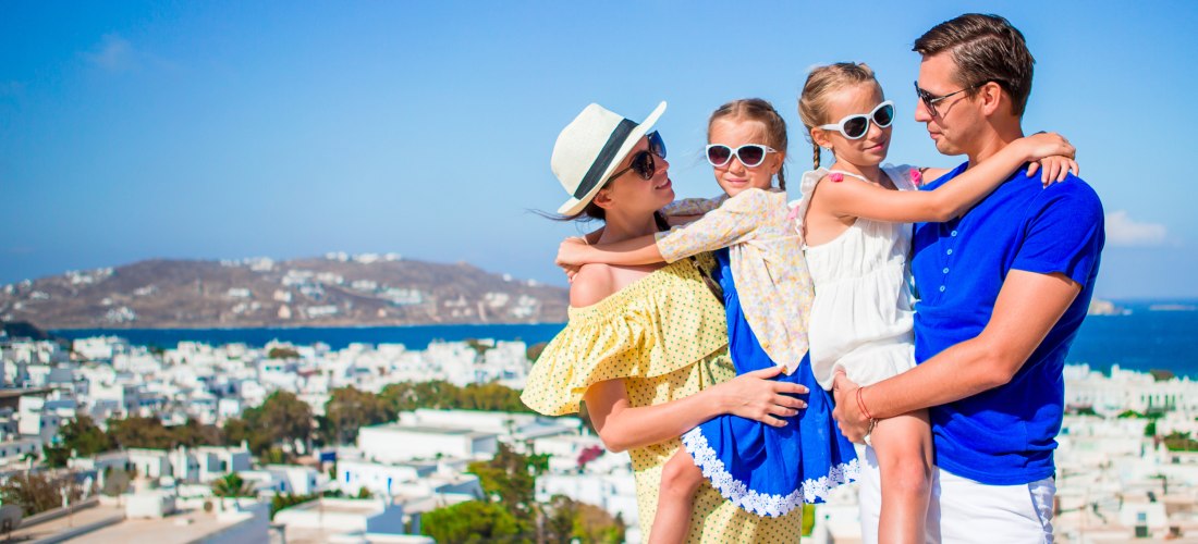 Fröhliche Familie mit zwei Kindern posiert auf einer Terrasse vor einer Küstenstadt mit weißen Häusern. Der Vater trägt ein blaues Shirt und hält eine Tochter auf dem Arm, die Mutter trägt ein gelbes Kleid und hält die andere Tochter. Alle tragen Sonnenbr