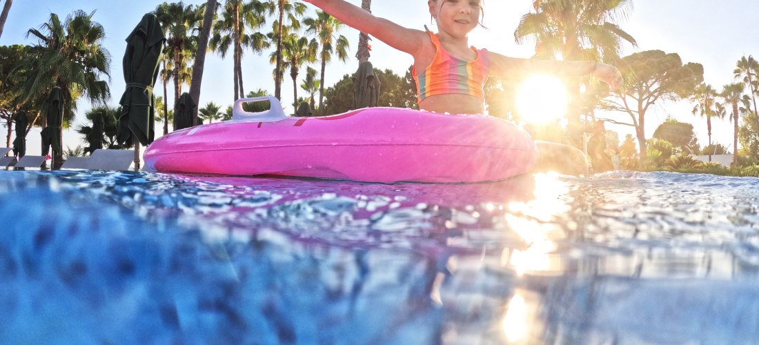 Lächelndes Kind auf pinker Luftmatratze im Pool, mit ausgestreckten Armen, im Hintergrund Palmen und tief stehende Sonne.