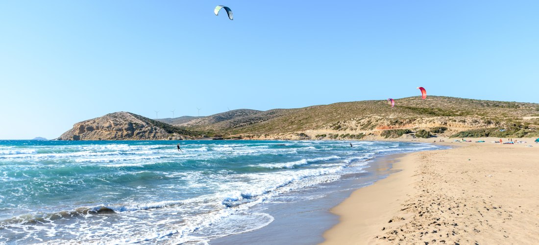 Weitläufiger Sandstrand mit sanften Wellen und klarem, türkisblauem Wasser. Am Himmel fliegen bunte Kites, während einige Kitesurfer über das Wasser gleiten. Im Hintergrund erstreckt sich eine hügelige, trockene Landschaft mit Windrädern auf den Bergrücke