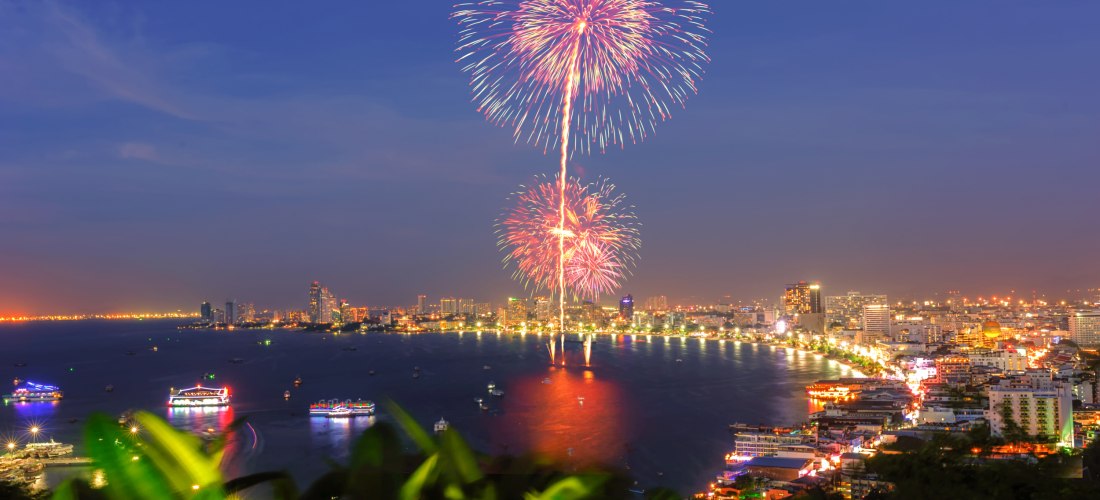 Farbenfrohes Feuerwerk über der Bucht von Pattaya bei Nacht. Im Vordergrund grüne Pflanzen, im Hintergrund leuchtende Boote auf dem Wasser und die hell erleuchtete Skyline der Stadt.