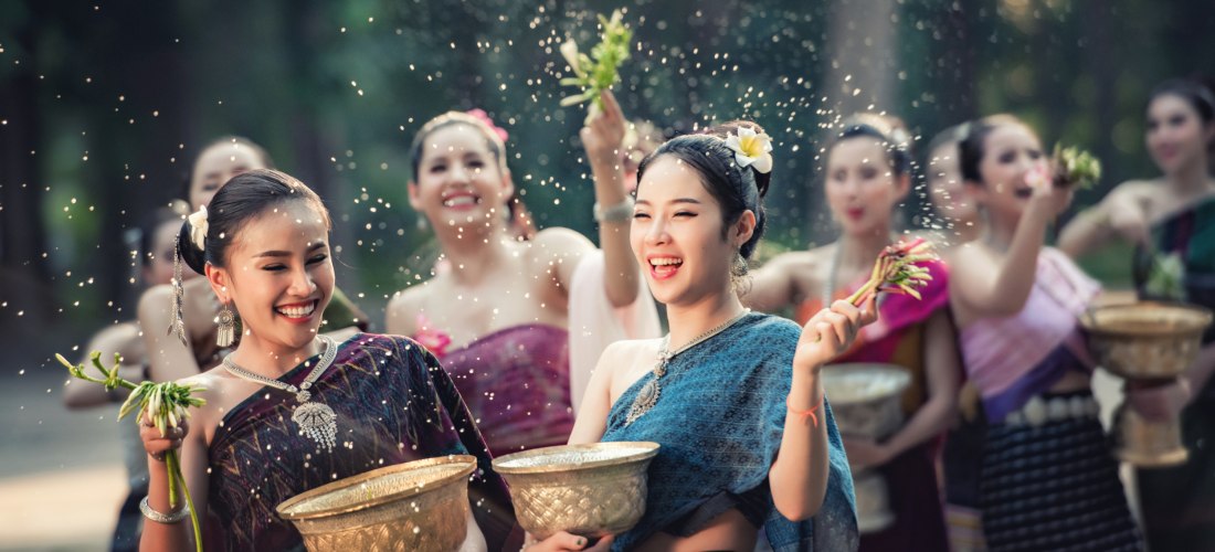 Mehrere thailändische Frauen in farbenfroher traditioneller Kleidung feiern das Songkran-Fest. Sie lachen, halten silberne Schalen und versprühen Wasser in der Luft.