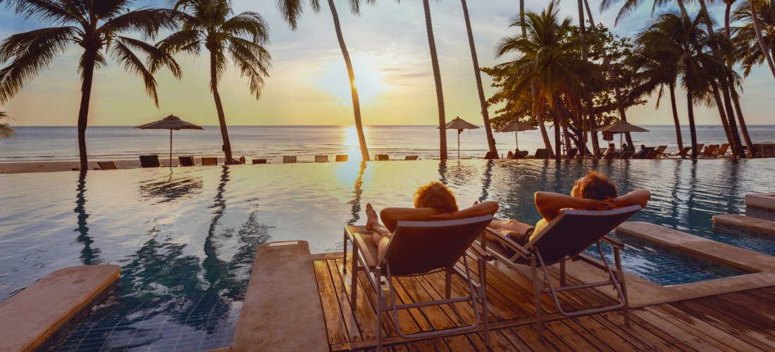 Ein Paar genießt den Sonnenuntergang in luxuriösen Liegestühlen am Infinity-Pool, umgeben von Palmen und mit Blick auf das ruhige Meer. Eine perfekte Szenerie für absolute Entspannung und Urlaubsfeeling.