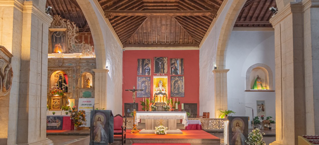 Blick in das Innere einer Kirche mit einem festlich geschmückten Altar, roten Teppichen, Heiligenfiguren und religiösen Gemälden. Das Gewölbe ist aus Holz, flankiert von steinernen Bögen.