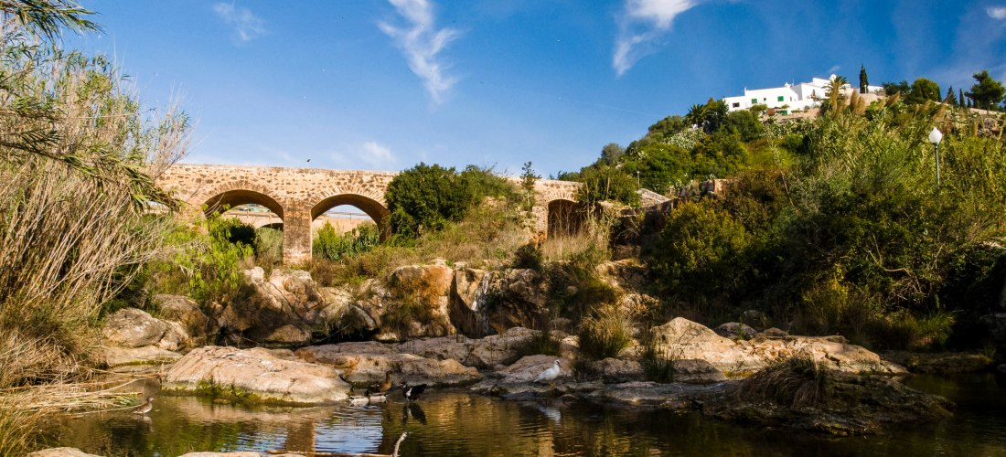 Historische Steinbrücke mit mehreren Bögen über einem flachen Flussbett, umgeben von Felsen, Sträuchern und Bäumen. Auf einem Hügel im Hintergrund steht ein weißes Haus vor blauem Himmel.